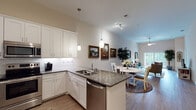 Located at 5500 Main St in New Port Richey, Florida, this single-floor residence offers a thoughtfully designed open-concept layout that seamlessly connects the kitchen, dining, and living areas. The kitchen is a standout feature, outfitted with crisp white shaker cabinetry, a subway tile backsplash, dark granite countertops, and a full suite of stainless steel appliances including a refrigerator, range, microwave, and dishwasher. A peninsula with bar seating and pendant lighting serves as both a functional workspace and a natural divider between the kitchen and dining area. The open living room benefits from soaring ceilings, light wood-style flooring, large windows that invite abundant natural light, and a ceiling fan. A cozy seating arrangement, framed artwork, and an indoor plant lend warmth to the space. The bedroom is a serene retreat featuring plush carpeting, multiple windows with blinds, and ample room for a dresser and nightstands. The bathroom is clean and well-finished, with a white vanity, stone-look countertop, tiled tub-shower combination, and neutral tile flooring. Throughout the residence, a cohesive neutral color palette and quality finishes create a comfortable and inviting atmosphere.