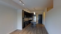 This single-floor space features a thoughtfully designed open-concept layout that seamlessly connects the kitchen and living areas. The kitchen is a standout element, outfitted with rich dark cabinetry that contrasts elegantly against light-toned walls and a classic subway tile backsplash. Stainless steel appliances, including a full-size refrigerator, built-in microwave, and dishwasher, provide a sleek, modern aesthetic. A freestanding kitchen island with a granite-style countertop offers additional prep space and casual seating, anchoring the kitchen within the open floor plan. Track lighting runs along the ceiling, providing targeted illumination throughout the kitchen, while a pair of pendant lights hangs above the island, adding a refined touch. The warm hardwood-style flooring extends throughout the main living area, lending continuity and a sense of warmth to the space. A dark front entry door with clean architectural framing provides a defined entryway, contributing to the unit's modern character. The neutral wall palette and efficient, well-organized layout create a functional yet stylish environment that is both inviting and easy to navigate. Overall, the space conveys a contemporary urban aesthetic with quality finishes throughout.
