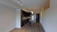 This single-floor space features a thoughtfully designed open-concept layout that seamlessly connects the kitchen and living areas. The kitchen is a standout element, outfitted with rich dark cabinetry that contrasts elegantly against light-toned walls and a classic subway tile backsplash. Stainless steel appliances, including a full-size refrigerator, built-in microwave, and dishwasher, provide a sleek, modern aesthetic. A freestanding kitchen island with a granite-style countertop offers additional prep space and casual seating, anchoring the kitchen within the open floor plan. Track lighting runs along the ceiling, providing targeted illumination throughout the kitchen, while a pair of pendant lights hangs above the island, adding a refined touch. The warm hardwood-style flooring extends throughout the main living area, lending continuity and a sense of warmth to the space. A dark front entry door with clean architectural framing provides a defined entryway, contributing to the unit's modern character. The neutral wall palette and efficient, well-organized layout create a functional yet stylish environment that is both inviting and easy to navigate. Overall, the space conveys a contemporary urban aesthetic with quality finishes throughout.