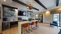 This single-floor amenity space blends modern industrial design with warm, inviting elements to create a versatile community lounge and business center. The open layout is anchored by bold exposed dark ceiling beams and light hardwood-style flooring that run throughout, lending the space a cohesive, contemporary character.

The kitchenette and lounge bar area serves as a central focal point, featuring dark cabinetry, a textured whitewashed brick backsplash, open shelving, and a wall-mounted flat-screen television. A generous white countertop island with reclaimed wood paneling offers seating for several guests via leather-upholstered bar stools, while a statement drum pendant light fixture adds dramatic overhead interest. The area also includes a sink, beverage refrigerator, and coffee station, making it well-suited for casual gatherings.

Adjacent to the kitchenette, the business center area is equipped with multiple computer workstations set along a built-in desk, illuminated by stylish industrial dome pendant lights. Large windows fitted with layered roller shades allow natural light to filter in, offering views of the outdoor pool area. Comfortable lounge seating — including bold blue and yellow accent chairs — is arranged nearby, creating a relaxed environment for informal meetings or downtime. The overall ambiance is polished yet approachable, balancing functionality with thoughtful design details throughout.