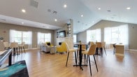 This well-appointed single-floor amenity space offers a dynamic and inviting environment designed for both relaxation and social engagement. The main lounge area features vaulted ceilings, warm hardwood-style flooring, and an abundance of natural light streaming through large windows dressed with sheer white curtains. Vibrant yellow accent chairs and a tufted gray sectional sofa are arranged around a sleek double-sided fireplace, above which a wall-mounted television is flanked by built-in open shelving. High-top bistro tables and bar-height seating provide flexible gathering options within the open-plan layout. A dedicated game room accommodates a full-size billiards table beneath modern pendant lighting, with French doors connecting it to the broader amenity space and a private phone booth or focus pod nearby. A café-style nook features a continuous navy velvet banquette set against yellow-accented built-in shelving, illuminated by geometric pendant lights and paired with round white tables and textured side chairs — ideal for casual work or conversation. The galley-style kitchen is outfitted with rich dark wood cabinetry, stainless steel appliances including a built-in microwave and dishwasher, light countertops, and a window that fills the space with natural light. Throughout, the cohesive design palette of warm neutrals, bold accents, and contemporary finishes creates an engaging and functional community environment.
