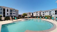 This outdoor pool area is designed for relaxation and recreation, offering a generously sized swimming pool with clear, turquoise water that reflects the open blue sky above. The pool's smooth, curved edges give it an inviting, resort-like aesthetic, while a smaller integrated spa or hot tub sits adjacent, providing a dedicated space for hydrotherapy and unwinding.

Surrounding the pool is a well-maintained deck finished in light-toned pavers, offering ample space for lounging. Teal-colored chaise lounge chairs and additional seating are arranged around the perimeter, providing plenty of options for sunbathing or socializing. Small side tables accompany the seating areas for added convenience.

A black wrought-iron fence encloses the pool area, ensuring both security and a clean visual boundary. Mature trees within and just beyond the enclosure offer pockets of natural shade. Signage is posted near the entrance, indicating pool rules and safety guidelines.

The backdrop features multi-story residential buildings with neutral-toned exteriors and pitched rooflines, contributing to a well-established community atmosphere. The entire space is bathed in abundant natural sunlight, creating a bright, welcoming environment ideal for outdoor leisure.