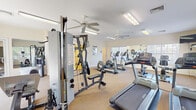 This single-floor fitness center is designed to support a well-rounded workout routine in a clean, functional environment. The main exercise area is generously proportioned, featuring wall-to-wall mirrors along one side that amplify the sense of space and allow users to monitor their form during workouts. Large windows fitted with blinds line the walls, flooding the room with natural light and offering views of the surrounding greenery.

The space is outfitted with a variety of cardio equipment, including multiple treadmills and elliptical machines, providing ample options for aerobic training. A multi-station cable weight machine anchors the strength training area, offering a range of resistance exercises. The flooring throughout the main workout area is a durable, cushioned carpet, providing comfort and stability underfoot. Two ceiling fans are mounted overhead, ensuring consistent air circulation throughout the room. Overhead fluorescent lighting supplements the natural light for visibility during any hour.

Adjacent to the main fitness floor are ADA-compliant restroom facilities. The restrooms feature warm-toned tile flooring, grab bars for accessibility, framed artwork on the walls, and clean, minimalist fixtures. Both men's and accessible facilities are clearly marked and well-maintained, contributing to an inclusive and user-friendly environment overall.