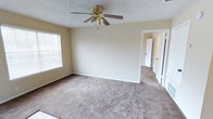 This single-story space presents a straightforward and functional layout with a clean, neutral aesthetic throughout. The room pictured features wall-to-wall carpeting in a warm beige tone, providing a soft and comfortable underfoot experience. Walls are painted in a light, neutral hue that contributes to a bright and open atmosphere. A gold-toned ceiling fan with wooden blades is centrally mounted on a textured popcorn ceiling, offering both air circulation and a modest decorative element. A window fitted with horizontal blinds allows natural light to filter into the space while maintaining privacy. White baseboards trim the perimeter of the room, adding a clean, finished look. A doorway visible in the background hints at additional rooms or hallway access, suggesting a connected and practical floor plan. The overall ambiance of the space is simple, uncluttered, and move-in ready, offering a blank canvas well-suited for a variety of furnishing styles and uses.
