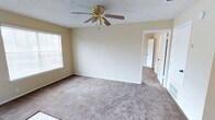 This single-story space presents a straightforward and functional layout with a clean, neutral aesthetic throughout. The room pictured features wall-to-wall carpeting in a warm beige tone, providing a soft and comfortable underfoot experience. Walls are painted in a light, neutral hue that contributes to a bright and open atmosphere. A gold-toned ceiling fan with wooden blades is centrally mounted on a textured popcorn ceiling, offering both air circulation and a modest decorative element. A window fitted with horizontal blinds allows natural light to filter into the space while maintaining privacy. White baseboards trim the perimeter of the room, adding a clean, finished look. A doorway visible in the background hints at additional rooms or hallway access, suggesting a connected and practical floor plan. The overall ambiance of the space is simple, uncluttered, and move-in ready, offering a blank canvas well-suited for a variety of furnishing styles and uses.