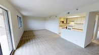 This single-floor space features an open-concept layout that seamlessly connects the living area to the kitchen. The spacious living area is finished with a light-toned patterned carpet, adding subtle texture to the neutral aesthetic. Smooth white walls and a vaulted ceiling contribute to an airy, open feel throughout the space.

The kitchen is accessible via a half-wall peninsula that defines the boundary between the two areas while maintaining visual openness. It features white cabinetry — both upper and lower — paired with a white range and a built-in sink beneath overhead fluorescent lighting. The cabinetry offers generous storage, and the layout provides practical countertop workspace. A dark wood accent panel beneath the upper cabinets adds a contrasting design detail.

HVAC vents are integrated into the ceiling, ensuring climate control throughout. The overall palette of whites and beiges creates a clean, cohesive environment that feels both functional and welcoming. The open floor plan makes the space feel larger than it might otherwise appear, lending itself well to flexible furniture arrangements and everyday living.