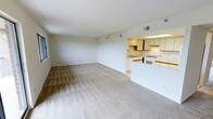 This single-floor space features an open-concept layout that seamlessly connects the living area to the kitchen. The spacious living area is finished with a light-toned patterned carpet, adding subtle texture to the neutral aesthetic. Smooth white walls and a vaulted ceiling contribute to an airy, open feel throughout the space.

The kitchen is accessible via a half-wall peninsula that defines the boundary between the two areas while maintaining visual openness. It features white cabinetry — both upper and lower — paired with a white range and a built-in sink beneath overhead fluorescent lighting. The cabinetry offers generous storage, and the layout provides practical countertop workspace. A dark wood accent panel beneath the upper cabinets adds a contrasting design detail.

HVAC vents are integrated into the ceiling, ensuring climate control throughout. The overall palette of whites and beiges creates a clean, cohesive environment that feels both functional and welcoming. The open floor plan makes the space feel larger than it might otherwise appear, lending itself well to flexible furniture arrangements and everyday living.