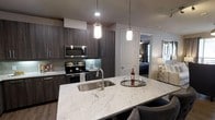 This single-floor residence showcases a thoughtfully designed open-concept layout that seamlessly connects the kitchen and living areas. The kitchen is a standout feature, centered around a generous island topped with striking white and grey veined quartz countertops that also house a deep stainless steel sink. Rich, dark wood-grain cabinetry lines the walls, providing ample storage while lending a contemporary and sophisticated aesthetic. Stainless steel appliances, including an electric range and over-the-range microwave, complement the modern design. Overhead, a pair of elegant cylindrical pendant lights cast a warm, focused glow over the island, enhancing the space's inviting ambiance. The kitchen flows naturally toward the rest of the residence, with a glimpse into what appears to be a well-appointed bedroom featuring soft, atmospheric lighting. Light-toned wood-style flooring runs throughout, tying the space together with a clean, cohesive look. The overall design balances dark cabinetry with lighter countertops and walls, creating a stylish contrast that feels both contemporary and refined. This space is well-suited for those who appreciate modern finishes and a functional, open living environment.