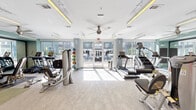 Located at 3027 Routh St in Dallas, TX, this fitness center is a thoughtfully designed, single-floor facility that offers a comprehensive workout environment. The space is bathed in natural light, courtesy of expansive floor-to-ceiling windows and glass doors that open to an outdoor area, creating an airy and energizing atmosphere throughout.

The layout is divided into distinct workout zones, each catering to different training needs. The cardio section is lined with a robust selection of equipment, including treadmills, a StairMaster, and rowing machines, all positioned to take advantage of the abundant natural light and outdoor views. Ceiling fans overhead contribute to a comfortable exercise environment.

Transitioning deeper into the space, a dedicated strength training zone features a multi-functional cable machine, weight benches, a barbell rack, and an assortment of free weights. A full-length mirror spans one wall, enhancing the sense of depth while allowing users to monitor their form.

A standout design feature is a textured dark mosaic accent wall that adds a touch of sophistication and visual contrast to the otherwise bright, neutral interior. Recessed linear lighting with warm amber accents along the ceiling provides an additional layer of ambiance, complementing the clean, modern aesthetic. The light-toned flooring with green carpet accents in the free weights area further zones the space. Overall, this fitness center is a well-appointed, modern facility suited for a full range of exercise routines.