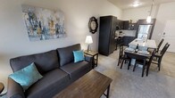 This single-floor unit features an efficient, open-concept layout that combines the living, dining, and kitchen areas into one cohesive space. The living area is anchored by a charcoal-toned sofa paired with a wooden coffee table, set atop neutral carpeting that extends throughout. A large abstract canvas in muted blues, yellows, and grays adds a decorative focal point to the main wall, complemented by a decorative wall clock that adds character to the space.

The kitchen runs along one wall and is outfitted with dark espresso-toned cabinetry, providing ample storage. Light countertops contrast the dark finishes, and the layout includes space for small appliances. A compact dining area with dark wood chairs sits adjacent to the kitchen, making it convenient for everyday meals.

The overall color palette is neutral and contemporary, with white walls and light flooring creating a clean, airy feel despite the compact footprint. The design prioritizes functionality, with each zone clearly defined yet visually connected, making the space feel organized and livable.