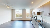 This single-floor unit presents a clean, modern design with a thoughtfully arranged open layout. The living area is a standout feature, with rich hardwood-style flooring and expansive windows that span a generous portion of the exterior wall, flooding the space with natural light and offering views of the surrounding greenery and cityscape. A ceiling fan adds both function and a finishing touch to the living space.

The unit includes two distinct kitchen configurations visible across the space. The primary kitchen is well-appointed with gray flat-front cabinetry, dark countertops, a classic white subway tile backsplash, and a full suite of stainless steel appliances including a range, microwave, dishwasher, and refrigerator. A secondary kitchen area features white cabinetry and similarly modern finishes, offering additional utility.

The bedrooms are carpeted and finished in neutral tones, providing a soft contrast to the hard flooring found elsewhere. The rooms are simply designed with clean white walls and recessed lighting, offering a versatile, move-in-ready environment.

The bathroom includes a full-size soaking tub with a clean white surround and chrome fixtures, maintaining the unit's consistent contemporary aesthetic throughout.