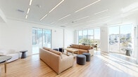 Located at 7000 Kiran Patel Dr in Winter Garden, FL, this contemporary amenity space offers a well-appointed, single-floor layout designed for both comfort and functionality. The open living and lounge area serves as the centerpiece of the space, featuring warm tan leather sofas, a wood-topped coffee table, and cylindrical ottomans arranged beneath a striking ceiling lined with sleek, linear LED lighting. Expansive floor-to-ceiling windows flood the area with abundant natural light and frame views of the surrounding community and palm-lined grounds. Light wood-look flooring runs throughout, contributing to the space's clean, modern aesthetic. The dining area provides counter-height seating along a window wall, complemented by stylish pendant lighting and continued views to the exterior. The kitchen is outfitted with ample white cabinetry, white quartz countertops, a deep sink, and distinctive floor-to-ceiling glazed tile walls in soft, neutral tones. A dedicated conference room features a large boardroom table, ergonomic executive chairs, a wall-mounted display screen, patterned carpet tiles, and full-height glazing offering views of the surrounding landscape, with glass partition walls lending the room a professional yet open character.
