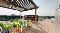 Located at 1178 Silas Deane Highway in Wethersfield, Connecticut, this expansive rooftop terrace offers a well-appointed outdoor amenity space designed for relaxation, dining, and socializing. The terrace is finished with large-format tiled flooring throughout and is bordered by sleek metal railings and lush green privacy hedging, creating a comfortable and inviting atmosphere open to the sky.

A standout architectural feature is the pergola structure positioned at one end of the terrace, providing partial shade over a vibrant lounge area furnished with bright green and white chairs accented by colorful patterned rugs. Adjacent to the pergola is a warm wood-clad privacy screen that adds a modern aesthetic element to the space.

A dedicated fire pit seating area anchors the central portion of the terrace, surrounded by green-cushioned chairs ideal for gathering. Nearby, a designated grilling zone features multiple full-sized gas grills alongside white dining tables and chairs, making it well-suited for outdoor meals and entertaining.

The remaining open areas of the terrace are furnished with chaise lounge chairs and additional dining sets, offering ample space for sunbathing and casual gatherings. Panoramic views of the surrounding Wethersfield landscape, including tree lines and neighboring structures, are visible from multiple vantage points across the terrace, enhancing the open-air experience.