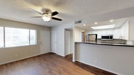 This single-story space offers an open, functional layout that seamlessly connects the living area to the kitchen. The living area is finished with rich, dark wood-style flooring that extends throughout, providing a warm and cohesive aesthetic. A ceiling fan with an integrated light fixture is centrally mounted on the textured ceiling, contributing to both comfort and ambiance. Recessed lighting adds supplemental illumination throughout.

The front entry features a classic paneled door with an adjacent window dressed with blinds, allowing natural light to filter in. Two interior doors are visible from the living space, offering access to additional rooms or storage areas.

The kitchen is positioned at the rear of the open layout and is elevated slightly by a raised breakfast bar with a dark countertop, creating a natural division between the living and cooking spaces. White cabinetry lines the upper walls, complementing the stainless steel refrigerator and built-in microwave. A skylight above the kitchen introduces additional natural light, brightening the cooking area. The overall palette of neutral tones and clean finishes gives the space a tidy, move-in-ready appeal.