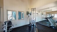This fitness room is a functional and well-appointed exercise facility designed to support a comprehensive workout routine. The space features notably high ceilings that create an open, airy atmosphere, preventing any sense of confinement during use. Smooth, light-colored walls are paired with dark, durable carpeted flooring, providing a clean and practical aesthetic throughout.

Natural light flows generously into the room through multiple large windows positioned along the walls, offering pleasant views of the surrounding outdoor landscaping, including shrubs and trees. A glass-paneled exterior door provides direct access to the outside, further enhancing the connection to the outdoors and allowing for easy ventilation.

The room is equipped with a multi-function cable strength training machine and True-brand treadmills, catering to both cardiovascular and resistance training needs. A weight bench is also present, adding versatility to the workout options available. Full-length mirrors line one wall, allowing users to monitor their form while exercising and visually expanding the sense of space within the room.

Posted exercise room guidelines and caution signage reflect a commitment to user safety and proper facility use. Overall, this fitness room combines practical equipment, abundant natural light, and a spacious design to create an inviting and effective environment for physical activity.