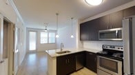 This single-floor, one-bedroom, one-bathroom unit offers a functional and modern open-concept layout that seamlessly connects the kitchen and living area. The kitchen serves as the centerpiece of the space, featuring dark espresso cabinetry that contrasts elegantly with light granite countertops. A peninsula island with an undermount sink provides additional prep space and casual seating potential, while pendant lighting above adds a stylish, contemporary touch. Stainless steel appliances, including a range, microwave, and dishwasher, complete the kitchen's polished look. A classic white subway tile backsplash ties the design together. The living area extends naturally from the kitchen, bathed in natural light from large windows with horizontal blinds. Light-toned hardwood-style flooring runs throughout, contributing to a warm and cohesive aesthetic. A ceiling fan in the living space adds both comfort and functionality. The overall ambiance is clean, modern, and efficiently designed, making excellent use of the available space while maintaining a bright and welcoming atmosphere throughout.