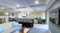 This single-floor space showcases a thoughtfully designed open-plan layout that seamlessly integrates a lounge, dining, and kitchen area into one cohesive environment. The architectural ceiling is a standout feature, with recessed coffered detailing and embedded can lighting that casts a clean, even glow across the space. Pendant lights hang above the kitchen zone, adding a layer of warmth and definition to the area.

The kitchen is well-appointed with stainless steel appliances, including a gas range, microwave, and a beverage refrigerator. A decorative mosaic tile backsplash in a scalloped pattern adds texture and visual interest to the cooking area. A wall-mounted flat-screen television is positioned conveniently for viewing from both the dining and lounge areas.

The dining bar features a dark countertop with ample seating provided by a row of upholstered high-back chairs, making it ideal for social gatherings. Adjacent to the kitchen and dining zone, the lounge area includes a light gray sofa with accent pillows and a wood-topped coffee table set over a plush area rug, creating a comfortable and inviting retreat. Light tile flooring ties the entire space together with a clean, contemporary aesthetic.