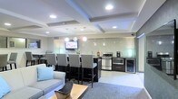 This single-floor space showcases a thoughtfully designed open-plan layout that seamlessly integrates a lounge, dining, and kitchen area into one cohesive environment. The architectural ceiling is a standout feature, with recessed coffered detailing and embedded can lighting that casts a clean, even glow across the space. Pendant lights hang above the kitchen zone, adding a layer of warmth and definition to the area.

The kitchen is well-appointed with stainless steel appliances, including a gas range, microwave, and a beverage refrigerator. A decorative mosaic tile backsplash in a scalloped pattern adds texture and visual interest to the cooking area. A wall-mounted flat-screen television is positioned conveniently for viewing from both the dining and lounge areas.

The dining bar features a dark countertop with ample seating provided by a row of upholstered high-back chairs, making it ideal for social gatherings. Adjacent to the kitchen and dining zone, the lounge area includes a light gray sofa with accent pillows and a wood-topped coffee table set over a plush area rug, creating a comfortable and inviting retreat. Light tile flooring ties the entire space together with a clean, contemporary aesthetic.