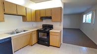 This single-floor space is laid out to offer practical, everyday functionality across its main areas. The kitchen is equipped with a black electric range and overhead exhaust hood, complemented by an L-shaped arrangement of light oak cabinetry that provides generous upper and lower storage. A built-in sink is positioned along the counter, and light-toned tile flooring runs throughout the kitchen area, contributing to a clean, utilitarian aesthetic.

Adjacent to the kitchen, the living area transitions to wall-to-wall carpeting in a neutral gray tone, creating a clear visual distinction between the two spaces. The living area is notably open, featuring white walls and a simple, unadorned ceiling that enhance the sense of available space. Multiple windows, including a wider multi-pane lattice-style window, allow natural light to filter into the room, brightening the interior. A wall-mounted electrical panel is visible in one corner of the living space, adding a practical element to the layout. Overall, the space presents a straightforward, functional design well-suited for comfortable everyday living.