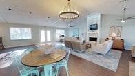 This single-floor space exudes a relaxed coastal charm, thoughtfully designed with a cohesive color palette of soft seafoam greens and crisp whites that create a light, airy atmosphere throughout. The open-concept layout seamlessly connects the dining and living areas, making the space feel expansive and inviting. Rich hardwood flooring runs throughout, grounding the space with warmth and texture. The dining area is anchored by a round wooden table surrounded by distinctive teal metal chairs, all beneath a striking circular chandelier that serves as a bold focal point. The living area is generously appointed with wicker and upholstered seating arranged around a classic white fireplace, complete with a mounted television above the mantle. A ceiling fan overhead ensures year-round comfort. A work or reception area is tucked near the back of the space, illuminated by natural light streaming through large windows and glass-paneled French doors that open to the outdoors. Decorative coastal accents and patterned area rugs tie the design together, while the textured ceiling adds architectural character. The overall ambiance is welcoming, bright, and thoughtfully curated — balancing function with a serene, beach-inspired aesthetic.