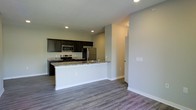 This single-floor space showcases a thoughtfully designed open-concept layout that seamlessly connects the kitchen and living area. The kitchen is a standout feature, equipped with stainless steel appliances — including a refrigerator, range, and microwave — complemented by dark espresso cabinetry that provides a bold contrast against the light grey walls. A peninsula with a granite-style countertop and an integrated sink serves as both a functional workspace and a natural divider between the kitchen and the adjoining living area, making it ideal for casual dining or entertaining. Recessed lighting throughout the kitchen ensures bright, even illumination. The flooring features wide-plank grey wood-style planks that run continuously through both spaces, lending a cohesive and contemporary feel. Neutral grey walls keep the interior bright and versatile. A doorway visible from the living area suggests additional rooms beyond, hinting at a functional floor plan. The overall aesthetic is clean, modern, and move-in ready, with a palette of dark, light, and neutral tones creating a well-balanced and inviting atmosphere.
