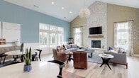 Located at 5744 Thorton St in Columbus, Ohio, this single-story community clubhouse blends modern design with warm, inviting aesthetics. The expansive main lounge is the heart of the space, showcasing soaring vaulted ceilings, soft blue-painted walls with crisp white wainscoting, and light wood-look tile flooring throughout. A dramatic floor-to-ceiling whitewashed stone fireplace serves as a striking focal point, complemented by a mounted flat-screen TV and a wood mantel. The lounge is bathed in natural light from large multi-pane windows, and an elegant chandelier with cascading strands adds a refined touch overhead. Comfortable seating arrangements — including leather club chairs, plush sofas, and layered area rugs — create a welcoming atmosphere ideal for gathering and relaxing. Adjacent to the lounge is a well-appointed kitchenette featuring dark cabinetry, white countertops, a sink, microwave, and beverage refrigerator. A dedicated co-working area offers a productive environment with workstations, high-top seating, tufted booth seating beneath brass pendant lights, and geometric wood-paneled accent walls. Sliding barn-style doors connect the work area to the main lounge, allowing for flexible use of the space. Throughout, thoughtful design details and curated furnishings create a cohesive, upscale communal environment.