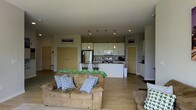 This single-floor space is designed around an open-concept layout that seamlessly integrates the living area and kitchen into one cohesive environment. The living area features light hardwood flooring and is anchored by a comfortable sectional sofa dressed with decorative pillows, creating a welcoming and casual atmosphere. Recessed lighting is installed throughout the ceiling, providing even, ambient illumination across the space.

The kitchen is positioned toward the rear and is outfitted with white cabinetry, pendant lighting hanging above the countertops, and stainless steel appliances including a refrigerator, microwave, and range. A kitchen island or breakfast bar provides additional counter space and seating with bar-height chairs, making it functional for both cooking and casual dining.

Neutral gray-toned walls complement the light flooring, giving the interior a clean, contemporary feel. Two doorways with golden-toned doors are visible along one wall, leading to additional areas of the unit. A smaller cabinetry alcove is also visible to one side, suggesting additional storage. Overall, the layout prioritizes an efficient, open flow ideal for everyday living.