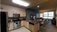 This single-floor unit showcases an open-concept layout that seamlessly connects the kitchen, dining, and living areas into one cohesive space. The kitchen is thoughtfully designed with light maple cabinetry providing ample storage, complemented by dark laminate countertops that add a grounding contrast. Stainless steel appliances, including an electric range, over-the-range microwave, and dishwasher, are fully integrated into the layout. A peninsula with an undermount double sink serves as a natural divider between the kitchen and the rest of the living space, offering additional prep and workspace. A bold slate-blue accent wall anchors the transition between the kitchen and the living area, adding depth and visual interest to the overall design. The living and dining area is warmly lit by a combination of recessed lighting and a floor lamp, creating an inviting ambiance. A window along the far wall allows natural light to filter in, brightening the space. The open layout and neutral color palette throughout make the unit feel cohesive, functional, and comfortable.