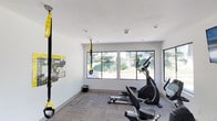 This compact yet functional fitness space is designed to support a well-rounded workout routine. The room features clean white walls and a neutral carpet floor, creating a bright and focused exercise environment. Large sliding windows line the exterior wall, flooding the space with natural light and offering pleasant views of the surrounding landscaping and greenery, making workouts feel less confined and more connected to the outdoors. The ceiling is finished with recessed lighting and ventilation, ensuring the space remains comfortable during use. Equipment includes an elliptical trainer, a stationary bike, and a rowing machine, providing a solid range of cardio options. A TRX suspension training system is mounted to the wall, offering versatile bodyweight resistance training. An informational exercise guide is posted on the wall nearby, serving as a handy reference for users. The overall design is practical and uncluttered, prioritizing functionality and ease of movement. Whether used for low-impact cardio, full-body suspension training, or a combination of both, this fitness room provides the essential tools needed for an effective workout in a clean, well-lit setting.