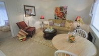 This single-floor residence offers a welcoming open-concept layout that connects the living and dining areas into one cohesive, functional space. The living area is anchored by a tan upholstered sofa adorned with patterned throw pillows, complemented by a plush burgundy accent chair and a dark round coffee table resting atop a gold and white area rug. Warm lighting from coordinating red-shaded lamps enhances the cozy ambiance, while bold floral wall art adds a vibrant, artistic focal point to the neutral walls. The light-toned carpet throughout contributes to the room's airy, comfortable feel. Transitioning naturally from the living space, the dining area features a round wooden table with white chairs, creating an intimate setting ideal for casual meals. Decorative accents, including a glass vessel filled with fruit, add a homey touch. The overall aesthetic balances warmth and functionality, with a color palette of golds, reds, and earth tones creating a cohesive and inviting atmosphere throughout the space.
