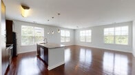 This single-floor residence offers a thoughtfully designed open-concept layout that connects the kitchen and living area seamlessly. Rich, polished hardwood flooring runs throughout the main living spaces, reflecting abundant natural light from multiple large windows that frame outdoor greenery views. The kitchen is a standout feature, anchored by a central island with granite countertops, an undermount sink, and pendant lighting overhead. Dark espresso cabinetry pairs with stainless steel appliances — including a range, microwave, and refrigerator — to create a sleek, modern aesthetic. The spacious living area extends generously from the kitchen, offering ample room for varied furniture arrangements.

A hallway connects the main living space to the private quarters, where a carpeted bedroom provides a comfortable retreat. The bedroom benefits from natural light through a dedicated window and includes access to a generously sized walk-in closet outfitted with wire shelving and hanging rods along multiple walls. The bathroom features a tub and shower combination with white tile surround accented by a neutral border tile, complemented by dark cabinetry with granite countertops and hardwood flooring. A louvered closet door in the hallway provides additional storage access.