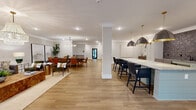 Located in Smyrna, GA, this thoughtfully designed clubhouse offers a generous, single-floor layout that seamlessly blends social and functional spaces. The heart of the clubhouse is a expansive open-plan area combining a fully equipped kitchen, dining space, and living room. The kitchen is anchored by a large island with quartz countertops and shiplap paneling, surrounded by navy bar-height seating. Oversized dark pendant lights with brass accents hang above, while a dramatic deep-toned tile backsplash and sage cabinetry add visual depth. Adjacent to the kitchen, the dining area accommodates a substantial table with leather chairs beneath a modern multi-bulb chandelier. The open living area flows naturally from this space, featuring wide-plank wood-look flooring and bright white walls throughout. A separate sitting lounge provides an intimate retreat, appointed with a cascading gold chandelier, curated artwork, and elegantly upholstered accent chairs arranged around a round cocktail table. A dedicated coworking alcove includes a built-in desk and accommodates two workstations. The entry foyer is bright and welcoming, framed by glass-paneled doors that draw in natural light and outdoor views. Crown molding and recessed lighting unify the interior with a clean, polished aesthetic throughout.