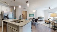 Located at 2604 Executive Dr in Venice, Florida, this single-story residence presents a thoughtfully designed open-concept floor plan that seamlessly connects the kitchen, dining, and living areas. The kitchen is a standout feature, showcasing an expansive granite-topped island with a double undermount sink and pendant lighting overhead. Cabinetry is finished in a two-tone combination of crisp white upper cabinets and sage-gray lower cabinets, complemented by a full suite of stainless steel appliances including a French door refrigerator, range, microwave, and dishwasher. Track lighting and additional pendant fixtures provide ample illumination throughout.

The dining area flows naturally from the kitchen and sits adjacent to a dedicated laundry room with full-size washer and dryer. The living room benefits from natural light through large windows and a glass door, with a ceiling fan adding comfort. Wood-look flooring runs throughout the main living areas, while the bedrooms feature plush carpet. Each bedroom includes a walk-in closet, and the bathroom presents white shaker-style cabinetry paired with granite countertops and wood-look tile flooring. The overall aesthetic is clean, contemporary, and consistently finished throughout.
