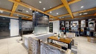This thoughtfully designed single-floor space makes a striking impression through its seamless blend of rustic and contemporary elements. The coffered ceiling, adorned with exposed natural wood beams and recessed lighting, anchors the space with warmth and architectural character. At the heart of the layout stands a dramatic multi-sided fireplace encased in a darkened metal surround with a glass base, serving as a compelling focal point that defines the open floor plan.

Large-format light-toned tile flooring flows throughout, creating a clean, cohesive foundation beneath the richly layered design. A reception or concierge area features a richly veined stone counter, adding a luxurious touch. Seating areas are carefully curated, including textured upholstered chairs around a wood dining table and intimate lounge nooks. One standout nook features a bold circular archway framing a banquette with leather seating and direct views to the exterior through floor-to-ceiling windows.

A cozy lounge alcove is set against a dramatic dark glazed brick accent wall, lit from above to highlight its texture, with a plush daybed and round wood side table. Built-in shelving and display cabinetry line the walls, adding functional elegance. Sliding barn-style doors and sculptural wall art contribute to the space's refined, urban-industrial aesthetic throughout.