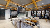 This thoughtfully designed single-floor space makes a striking impression through its seamless blend of rustic and contemporary elements. The coffered ceiling, adorned with exposed natural wood beams and recessed lighting, anchors the space with warmth and architectural character. At the heart of the layout stands a dramatic multi-sided fireplace encased in a darkened metal surround with a glass base, serving as a compelling focal point that defines the open floor plan.

Large-format light-toned tile flooring flows throughout, creating a clean, cohesive foundation beneath the richly layered design. A reception or concierge area features a richly veined stone counter, adding a luxurious touch. Seating areas are carefully curated, including textured upholstered chairs around a wood dining table and intimate lounge nooks. One standout nook features a bold circular archway framing a banquette with leather seating and direct views to the exterior through floor-to-ceiling windows.

A cozy lounge alcove is set against a dramatic dark glazed brick accent wall, lit from above to highlight its texture, with a plush daybed and round wood side table. Built-in shelving and display cabinetry line the walls, adding functional elegance. Sliding barn-style doors and sculptural wall art contribute to the space's refined, urban-industrial aesthetic throughout.