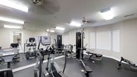 This fitness center is designed to support a well-rounded workout routine in a functional and well-maintained environment. The space is outfitted with a selection of cardio equipment, including a True elliptical trainer and a treadmill, alongside multi-station strength training machines that cater to a range of exercise needs. Floor-to-ceiling mirrors line the walls, creating a sense of openness and allowing users to monitor their form during workouts. The flooring is dark and durable, appropriate for a high-traffic fitness setting. Natural light filters in through multiple windows fitted with white blinds, complementing the overhead fluorescent lighting to keep the space bright and well-lit. Ceiling fans are installed throughout to maintain air circulation and comfort during exercise. The overall layout is organized and spacious, providing ample room to move between equipment. This single-floor fitness facility offers a practical and fully equipped space for both cardiovascular and strength-based training.