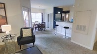 This single-floor residence showcases a thoughtfully designed open-concept layout that integrates the living, dining, and kitchen areas into one cohesive, flowing space. Plush wall-to-wall carpeting runs throughout the main living and dining areas, adding warmth and comfort underfoot. The living area provides a welcoming atmosphere with neutral tones and ample room for furnishings, while large sliding glass doors and windows flood the space with abundant natural light, creating an airy and inviting ambiance. The dining area comfortably accommodates a full dining set beneath an elegant pendant light fixture, making it ideal for everyday meals or entertaining. A bold navy blue accent wall serves as a striking focal point, adding a pop of color and visual depth to the open layout. The kitchen is accessible via a breakfast bar counter with seating for bar stools, offering a casual dining option and a natural gathering space. Track lighting and a pass-through design keep the kitchen connected to the living areas while maintaining a sense of defined space. Overall, this residence combines functional design with modern aesthetic touches, creating a bright and comfortable environment suited for a variety of lifestyles.
