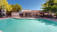 Located at 3135 Comanche Rd NE in Albuquerque, New Mexico, this outdoor amenity space showcases a generously sized, freeform swimming pool as its central feature. The pool's clear turquoise water is complemented by a wide concrete pool deck that provides ample room for lounging and socializing. Scattered throughout the deck are vibrant orange lounge chairs and coordinating patio furniture, including dining sets that invite residents to relax and gather under the New Mexico sun.

The space is framed by a lush canopy of mature deciduous trees, offering natural shade and a refreshing contrast to the warm desert climate. A wrought-iron perimeter fence encloses the area, providing both security and a sense of definition within the community grounds.

Adjacent to the pool, a single-story clubhouse building with a distinctive red-tiled roof and covered pergola-style porch serves as an architectural anchor. Large windows and glass doors along the clubhouse facade suggest an interior gathering space that opens directly to the pool deck. The surrounding multi-story residential buildings, finished in neutral stucco tones, frame the courtyard and reinforce the cohesive Southwestern architectural style. Under Albuquerque's abundant blue skies, this pool area offers a bright, welcoming outdoor environment year-round.