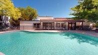 Located at 3135 Comanche Rd NE in Albuquerque, New Mexico, this outdoor amenity space showcases a generously sized, freeform swimming pool as its central feature. The pool's clear turquoise water is complemented by a wide concrete pool deck that provides ample room for lounging and socializing. Scattered throughout the deck are vibrant orange lounge chairs and coordinating patio furniture, including dining sets that invite residents to relax and gather under the New Mexico sun.

The space is framed by a lush canopy of mature deciduous trees, offering natural shade and a refreshing contrast to the warm desert climate. A wrought-iron perimeter fence encloses the area, providing both security and a sense of definition within the community grounds.

Adjacent to the pool, a single-story clubhouse building with a distinctive red-tiled roof and covered pergola-style porch serves as an architectural anchor. Large windows and glass doors along the clubhouse facade suggest an interior gathering space that opens directly to the pool deck. The surrounding multi-story residential buildings, finished in neutral stucco tones, frame the courtyard and reinforce the cohesive Southwestern architectural style. Under Albuquerque's abundant blue skies, this pool area offers a bright, welcoming outdoor environment year-round.