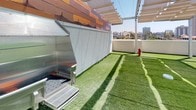 Located at 550 W Date St in the Little Italy neighborhood of San Diego, this rooftop pet amenity space is a thoughtfully designed outdoor area that caters to both pets and their owners. The level is covered in lush artificial turf, providing a clean, low-maintenance surface ideal for pets to play and roam freely. A dedicated stainless steel pet wash station with a flexible hose makes post-walk cleanups convenient and efficient. Overhead, a pergola-style shade structure with sail canopies offers welcome relief from the Southern California sun, while string lights add a pleasant ambiance for evening visits. Vibrant red lounge chairs are positioned throughout the space, inviting residents to relax while their pets enjoy the area. A fire hydrant fixture serves as a charming and functional accent. Sturdy metal railings enclose the perimeter, ensuring safety while maintaining open sightlines. Striking warm-toned architectural cladding adorns the surrounding building facade, adding visual character. The space offers impressive panoramic views of the San Diego downtown skyline, making it a standout amenity that combines practicality with an elevated outdoor experience in the heart of the city.