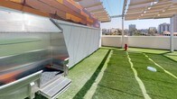 Located at 550 W Date St in the Little Italy neighborhood of San Diego, this rooftop pet amenity space is a thoughtfully designed outdoor area that caters to both pets and their owners. The level is covered in lush artificial turf, providing a clean, low-maintenance surface ideal for pets to play and roam freely. A dedicated stainless steel pet wash station with a flexible hose makes post-walk cleanups convenient and efficient. Overhead, a pergola-style shade structure with sail canopies offers welcome relief from the Southern California sun, while string lights add a pleasant ambiance for evening visits. Vibrant red lounge chairs are positioned throughout the space, inviting residents to relax while their pets enjoy the area. A fire hydrant fixture serves as a charming and functional accent. Sturdy metal railings enclose the perimeter, ensuring safety while maintaining open sightlines. Striking warm-toned architectural cladding adorns the surrounding building facade, adding visual character. The space offers impressive panoramic views of the San Diego downtown skyline, making it a standout amenity that combines practicality with an elevated outdoor experience in the heart of the city.
