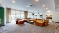 Located at 210 S Main St in Kannapolis, NC, this single-floor community amenity space is thoughtfully designed to foster relaxation and social interaction. The centerpiece of the lounge is an expansive sectional sofa in rich cognac leather, arranged around a low-profile coffee table atop a textured, patterned carpet that adds warmth and visual depth to the room. Large floor-to-ceiling windows fitted with sheer roller shades and floor-length drapes flood the space with natural light while offering views of the outdoor pool and surrounding community.

A dedicated media area features a large wall-mounted flat-screen television set against a bold teal accent wall, complemented by a wood-tone credenza with decorative accents. Adjacent to this is a rustic, wood-paneled accent wall that adds an organic, textural contrast to the otherwise contemporary palette. Small bistro-style tables with modern chairs provide intimate seating nooks ideal for casual conversation or individual use.

A separate dining or meeting alcove showcases a marble-top oval table paired with sculpted cream chairs, set against a weathered, tile-textured feature wall with curated artwork. Recessed lighting and pendant fixtures throughout ensure consistent, well-distributed illumination. The overall ambiance is modern yet inviting, balancing sophisticated design elements with comfortable, functional furnishings.