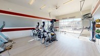 This fitness center is a thoughtfully designed single-floor facility that balances functionality with an inviting aesthetic. The space is defined by its crisp white walls adorned with a bold red horizontal stripe that wraps continuously throughout, creating a energetic and cohesive visual identity. Light gray flooring runs throughout, providing a clean, modern foundation underfoot.

Natural light is a standout feature, with expansive floor-to-ceiling windows and a large glass garage-style door along one wall, offering open outdoor views and flooding the interior with sunlight. The elevated ceilings, equipped with a ceiling fan and rows of fluorescent lighting, enhance the sense of openness and ensure the space remains well-lit at all times.

The cardio zone is well-appointed, featuring multiple treadmills, elliptical machines, and a stationary bike, with several units equipped with personal screens. A functional training area includes a cable machine, a SmartMat workout guide station, and a dedicated accessory station with towels, fitness balls, and a work surface. Wall-mounted mirrors and a flat-screen TV round out the amenities, making this a comprehensive and comfortable fitness environment.