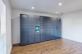 Located at 6075 Grelot Rd in Mobile, Alabama, this single-floor package locker room is designed to provide a streamlined and secure parcel retrieval experience. The space is defined by its clean, minimalist aesthetic, featuring crisp white walls, recessed ceiling lighting, and warm wood-look flooring that runs throughout the room, creating a bright and welcoming environment.

The centerpiece of the space is a large-scale Amazon Hub smart locker system, which spans nearly the full length of one wall. The unit offers an extensive array of individual compartments in varying sizes, accommodating packages of different dimensions. A central touchscreen interface allows for easy, self-service package pickup. The locker system's slate-blue finish provides a sleek, modern contrast against the white walls.

Natural light enters the room through a full-lite glass-panel entry door, offering a view of the surrounding tree-lined outdoor area. A rustic wood and metal console table near the entrance adds a functional accent, complementing the overall design. A security camera is also visible, underscoring the room's attention to safety and monitoring. The open floor plan allows for comfortable movement throughout the space, making package retrieval efficient and hassle-free.