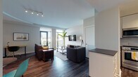 The Peach Studio is a thoughtfully designed one-bedroom space located in Atlanta, Georgia, offering a clean, modern aesthetic across a single floor. The open-concept living and kitchen area serves as the heart of the unit, where dark hardwood-style flooring contrasts elegantly with crisp white walls and ceilings. Floor-to-ceiling windows flood the living room with natural light and provide direct access to a private balcony, offering outdoor views of the surrounding Atlanta cityscape.

The kitchen features white gloss cabinetry extending to the ceiling, a subway tile backsplash, stainless steel appliances including a full-size refrigerator and dishwasher, and a dark-topped peninsula that defines the space. Track lighting and a bold black entry door add modern character near the entrance.

The living area accommodates comfortable seating, a wall-mounted flat-screen TV, and a warm wood media console. The bedroom is a serene retreat, furnished with a king-size bed, a bold upholstered headboard, matching nightstands, a full-length mirror, and a dedicated workspace with a desk and chair.

The bathroom is finished with neutral tile flooring, white cabinetry, a dark countertop, a large mirror, and a soaking tub with marble-style tile surround, completing this well-appointed Atlanta residence.