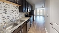 This single-floor studio is thoughtfully designed to maximize functionality within a compact, open footprint. Wood-style plank flooring flows throughout the main living and kitchen areas, contributing to a warm, cohesive aesthetic. The living area is anchored by a ceiling fan and benefits from generous natural light, delivered through a set of French doors that open onto a private balcony with metal railing, offering outdoor views of the surrounding area.

The kitchen runs along one wall in a galley-style configuration, featuring dark espresso cabinetry with brushed nickel hardware, granite-look countertops, a stainless steel double-basin sink, and a mosaic tile backsplash in neutral tones. Stainless steel appliances—including a refrigerator, electric range, built-in microwave, and dishwasher—complete the kitchen's functional setup.

Adjacent to the main entry, a double-door closet provides convenient storage. A hallway connects the living space to the full bathroom, which includes a tub-shower combination with tile surround, a dark-cabinet vanity with granite-look countertop, tile flooring, and brushed nickel fixtures throughout. The overall palette of soft grays and warm neutrals lends the space a clean, modern character.