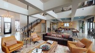 Located at 5260 Franklin St in Hilliard, Ohio, this two-story community space blends industrial-modern design with warm, inviting details throughout. The ground floor welcomes visitors through a bright, airy entry foyer with soaring ceilings, polished concrete floors, and expansive black-framed windows that flood the space with natural light and offer views of the surrounding neighborhood. The main lounge area is generously proportioned, featuring a dramatic double-height open ceiling, a mix of leather seating, a tufted sectional sofa, and a rustic wood coffee table anchored by a colorful area rug. A metal staircase with black iron railings leads to the upper level. Adjacent to the lounge, a well-appointed kitchen and bar area showcases shiplap wood-paneled walls, teal-accented ceiling details, dark stone countertops, pendant lighting, and bar-height seating. A dedicated business center offers workstation terminals with shiplap backdrops and bench seating. The upper level features hardwood flooring, a bright lounge with floor-to-ceiling windows overlooking the street, high-top communal tables, and a branded feature wall. Throughout, the design balances reclaimed wood textures, polished surfaces, and curated lighting to create a cohesive, modern-rustic ambiance.