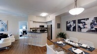 This single-floor space showcases an open-concept layout that seamlessly connects the kitchen, dining, and living areas. The kitchen is equipped with stainless steel appliances, including a refrigerator, stove, and microwave, complemented by white cabinetry and dark countertops. A breakfast bar with sleek acrylic barstools provides additional seating and acts as a natural divider between the kitchen and dining space. The dining area accommodates a full-sized wooden table with modern white chairs, set beneath a stylish pendant light that adds a warm focal point to the room. Warm hardwood-style flooring runs throughout the space, adding cohesion and a welcoming feel. The open sightlines extend into a bright bedroom area, visible in the background, suggesting a well-connected and functional floor plan. Neutral-toned walls keep the space feeling clean and versatile, while decorative touches such as a bold framed artwork piece and indoor greenery add personality. The overall ambiance is contemporary and well-appointed, balancing practicality with modern aesthetic sensibility.