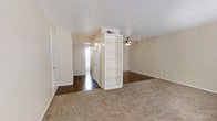 This single-story space presents an open, functional layout with a neutral color palette throughout. The main living area is finished with plush carpet flooring, creating a comfortable underfoot feel, while an adjoining hallway and adjacent rooms feature dark hardwood-style flooring that adds contrast and visual interest.

A freestanding white bookshelf serves as a partial room divider, providing subtle separation between the living and transitional areas without fully closing off the space. This detail adds both practical storage and a degree of architectural definition to the open floor plan.

The living area is equipped with a ceiling fan fitted with a light fixture, offering both ventilation and ambient lighting. Walls are finished in a warm off-white tone, complemented by a textured popcorn ceiling that is consistent throughout the space.

A hallway extends toward the back of the unit, where natural light filters in through what appears to be a window or exterior door, brightening the corridor. Floor vents are visible, indicating a central HVAC system. The overall ambiance is clean, neutral, and move-in ready, offering a versatile canvas for a variety of furnishing arrangements.