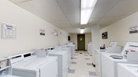 This single-floor shared facility offers two distinct functional areas designed to enhance community living. The laundry room is generously sized, lined with rows of commercial-grade white washing machines and dryers along both sides of the space. Bright fluorescent lighting illuminates the white drop-ceiling tiles and light vinyl flooring with gray accents, creating a clean, efficient environment. A posted "Last Wash" sign and clearly marked walls keep the space organized and user-friendly.

The community lounge is a warm and inviting gathering space with white shiplap-style wall paneling and a white paneled ceiling fitted with recessed lighting. Comfortable seating includes a plush navy sofa, coordinating armchairs with floral upholstery, and a round wood-top coffee table. Natural light filters in through horizontal windows positioned along the upper walls. A bookshelf stocked with reading materials, a ping-pong table, and a media console with a built-in electric fireplace add recreational variety. A kitchenette area along one wall features warm wood cabinetry, a granite-style countertop, a sink, a mini-refrigerator, and a microwave, making the lounge a fully functional and comfortable communal retreat.